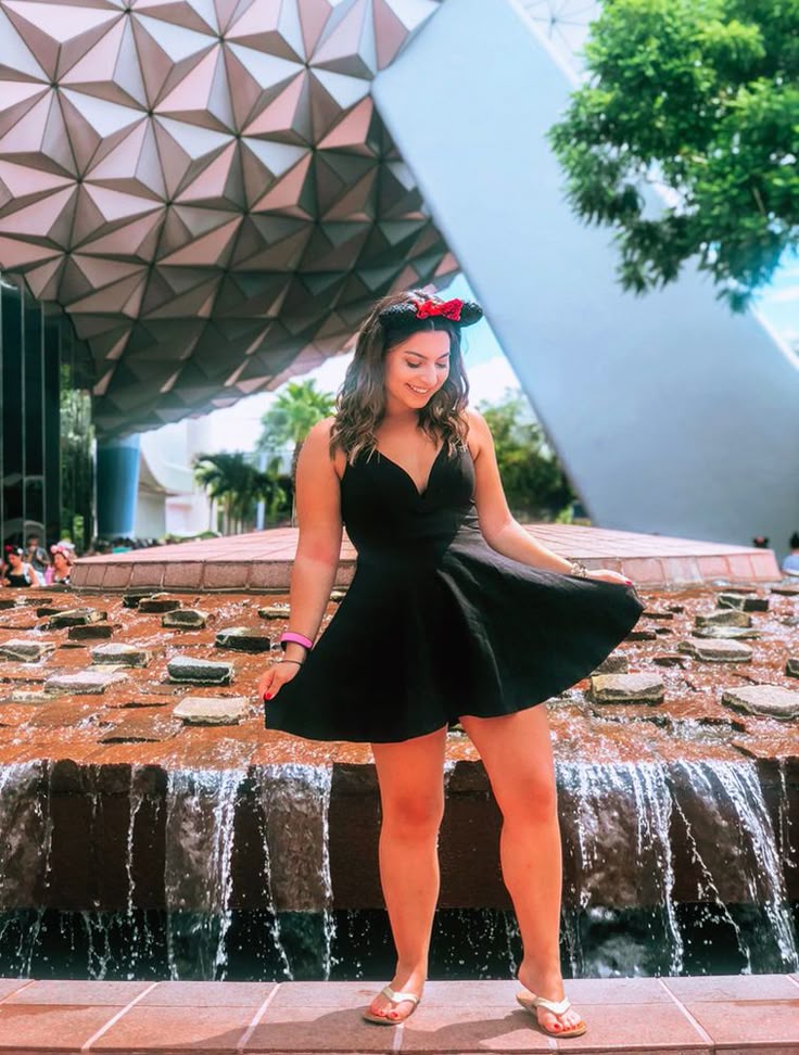 This may contain: a woman standing in front of a fountain wearing a black dress with a red bow on her head
