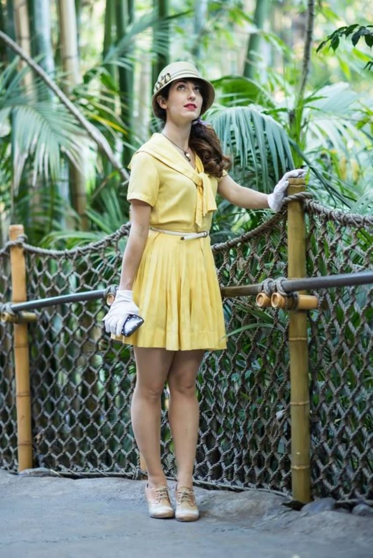 This may contain: a woman in a yellow dress and hat is standing on a bridge with palm trees behind her